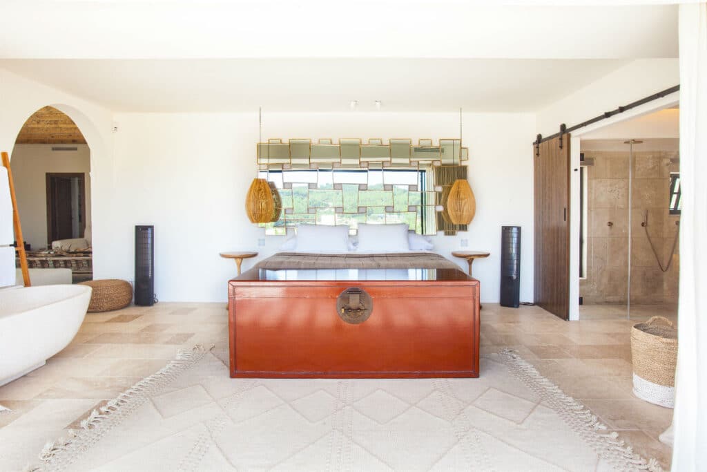 Bright, minimalist bedroom with a wooden bed frame, white bedding, large decorative mirror, and woven lamps exudes luxury travel vibes. A sliding barn door opens to an en-suite bathroom while a neutral area rug covers the light stone tile flooring. David Pearson Travel