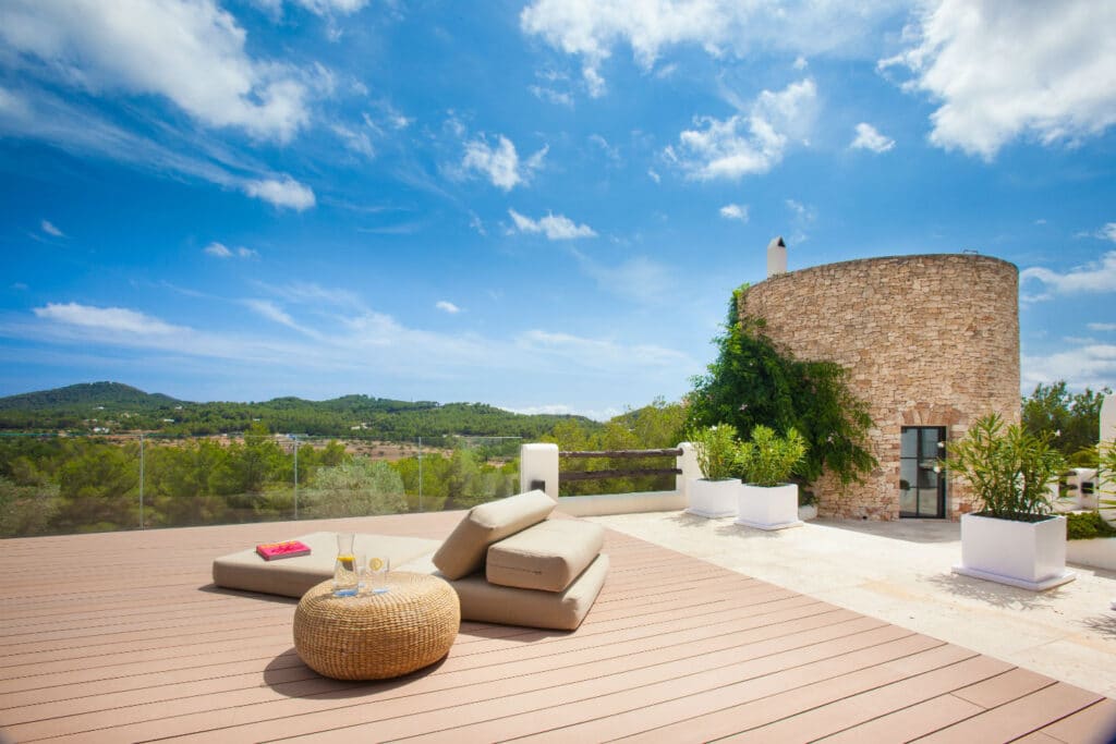 A sunlit terrace with plush cushions and a wicker table on wooden decking evokes the essence of luxury travel. To the right, a cylindrical stone tower enveloped in greenery adds charm. In the distance, rolling hills and a clear blue sky complete the serene setting. David Pearson Travel