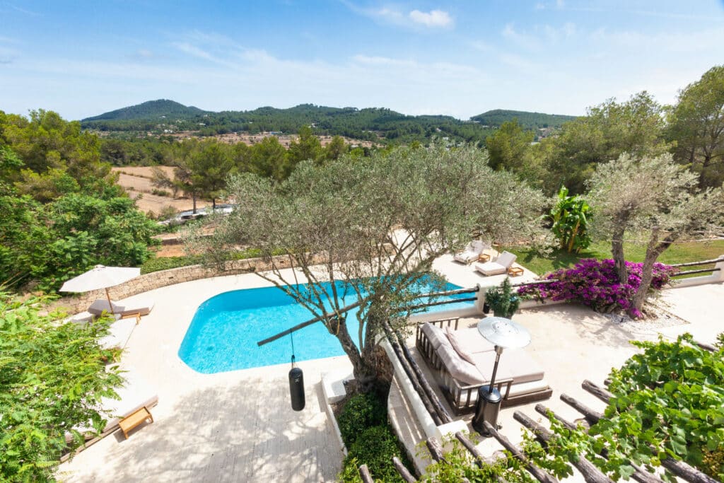 A serene outdoor scene perfect for luxury travel, featuring a swimming pool surrounded by lounge chairs and umbrellas. A large tree and vibrant purple flowers adorn the poolside, set against a backdrop of rolling hills and a clear blue sky. David Pearson Travel