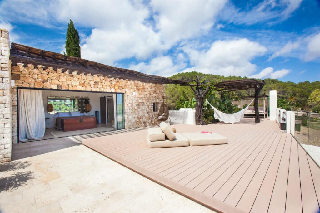 Spacious patio with wooden decking featuring beige lounge cushions and two white hammocks under shaded pergolas. Experience luxury travel adjacent to a stone building with large sliding glass doors, revealing an elegant interior living area. Lush greenery and blue sky in the background. David Pearson Travel