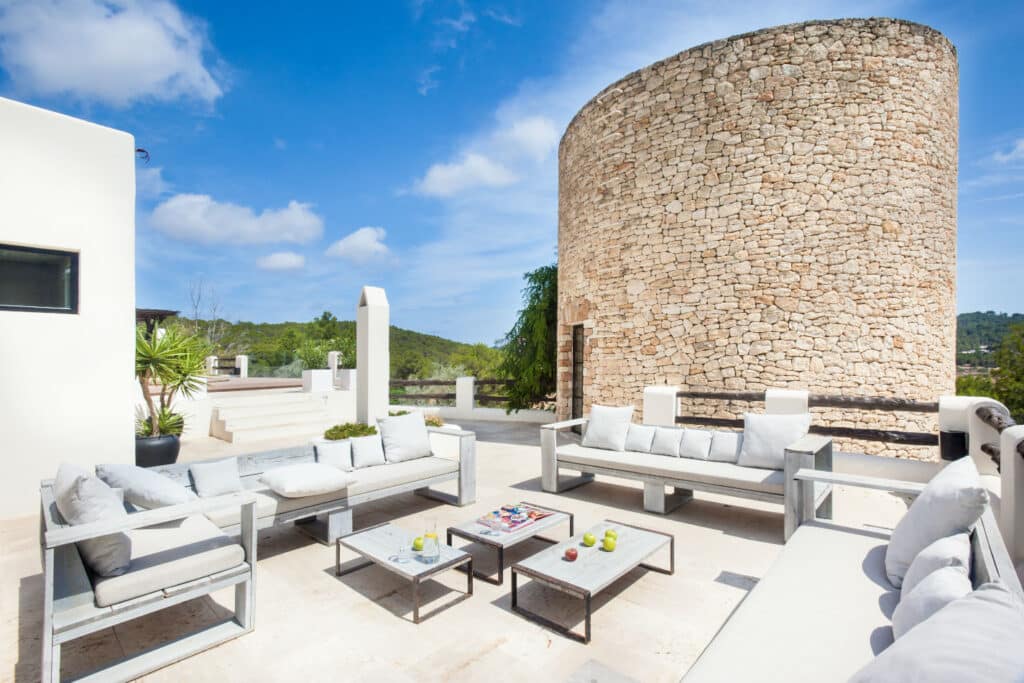 Experience the essence of luxury travel on this outdoor patio, featuring white sofas and a table nestled on a stone terrace. A grand round stone wall stands majestically in the background, with potted plants and trees framing breathtaking views of lush green hills under a crystal-clear sky. David Pearson Travel