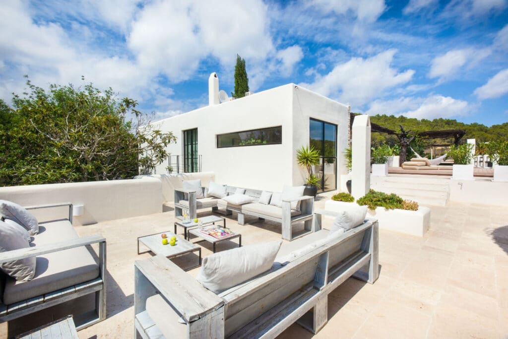 Modern white villa with a spacious rooftop terrace featuring cushioned wooden seating, a coffee table, and assorted plants. Surrounded by greenery and under a partly cloudy blue sky, it offers the perfect setting for luxury travel seekers looking to unwind in style. David Pearson Travel