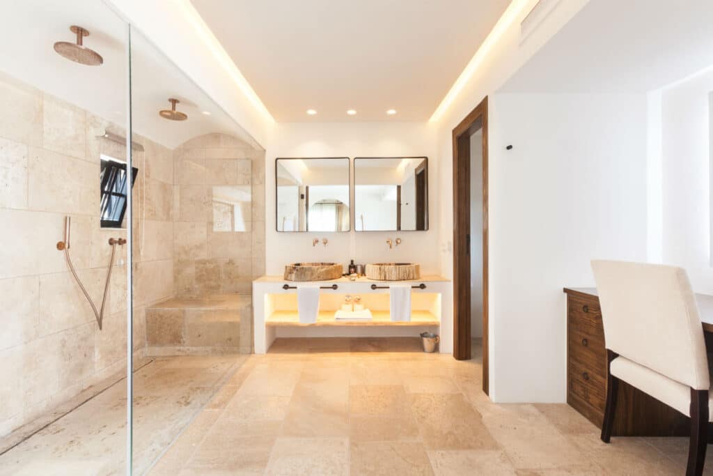 Experience a modern bathroom reminiscent of luxury travel, with beige stone tiles and a spacious glass-enclosed shower. It boasts a double vanity adorned with round vessel sinks, mirrors, and a wooden desk paired with a white chair. Recessed lighting adds an elegant touch to the serene atmosphere. David Pearson Travel