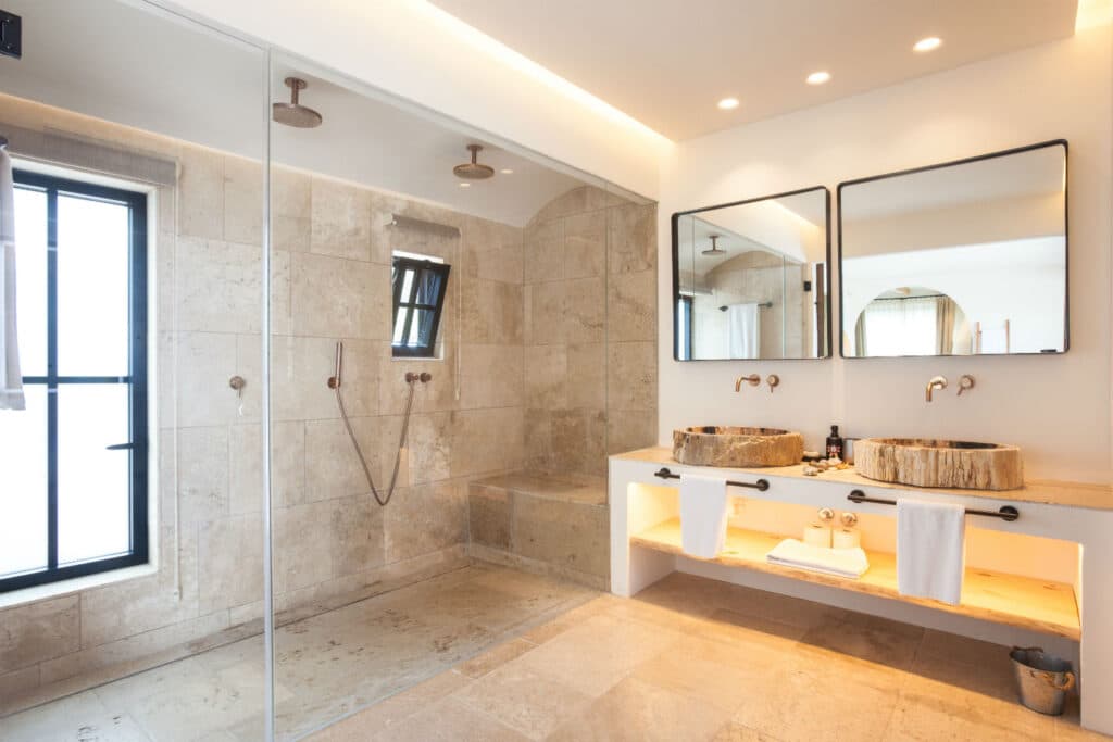 Spacious modern bathroom reminiscent of luxury travel, featuring a large walk-in shower with dual rainfall showerheads and beige tiles. A double vanity with vessel sinks, two mirrors, and neatly arranged towels on an open shelf is on the right. Natural light enters through a window. David Pearson Travel