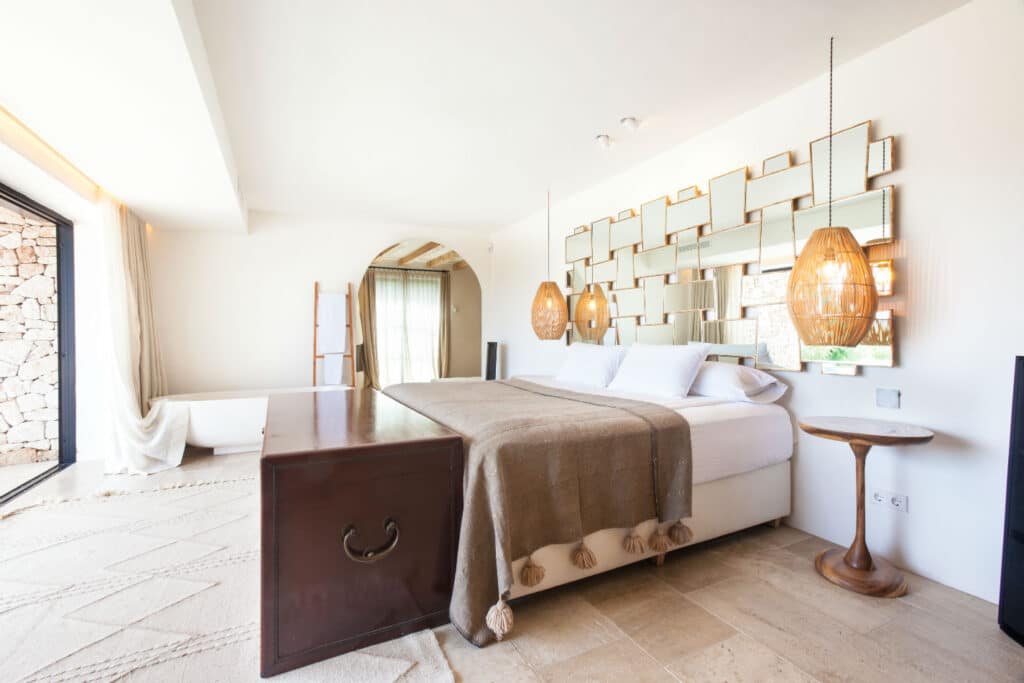 Modern bedroom with a large bed draped in a brown blanket, exuding a touch of luxury travel. Its framed by a geometric mirrored wall and two hanging woven lamps. A wooden chest sits at the beds foot, next to a narrow round table. A large window welcomes abundant natural light inside. David Pearson Travel
