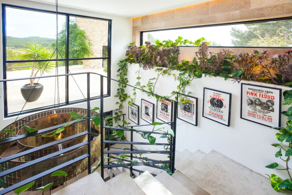Modern staircase with metal railings, adorned with vibrant green plants and framed Pink Floyd posters on the wall—evoking an air of luxury travel. Large windows invite natural light, highlighting a potted plant visible outside on the sunlit patio. David Pearson Travel