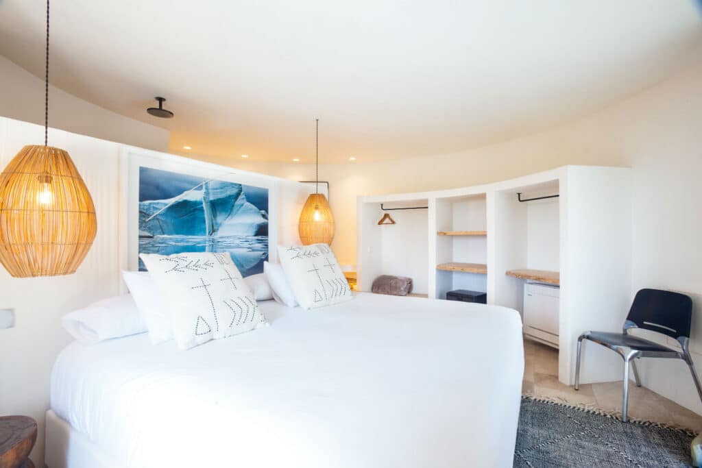 A modern bedroom exuding luxury travel vibes features a large bed with white linens and decorative pillows. Two woven pendant lights hang over the bed. A photograph of a glacier graces the wall, while open shelves and a chair in the corner complete the rooms clean, minimal design. David Pearson Travel