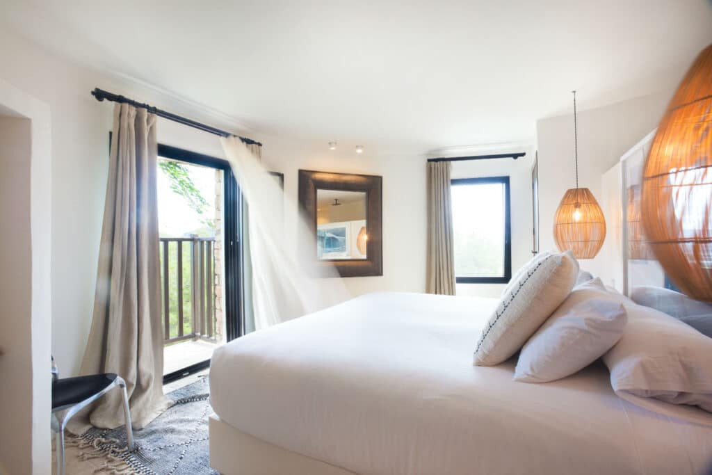 Bright, airy bedroom with a large bed, white bedding, and decorative pillows—perfect for luxury travel relaxation. Two pendant lights hang on the right. Curtains frame two windows, one leading to a small balcony with greenery outside. A mirror is mounted on the wall opposite the bed. David Pearson Travel