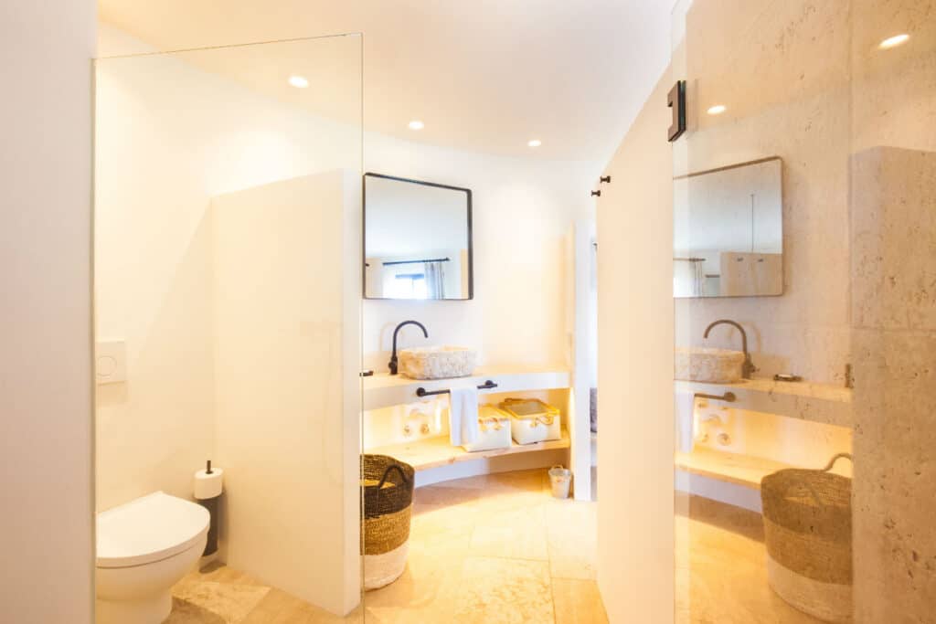 A modern bathroom with a glass shower, white toilet, and dual sinks evokes the elegance of luxury travel. Light beige walls, a large mirror above the sinks, and wicker baskets under the counter add to its charm. Recessed lighting enhances this tranquil oasis. David Pearson Travel