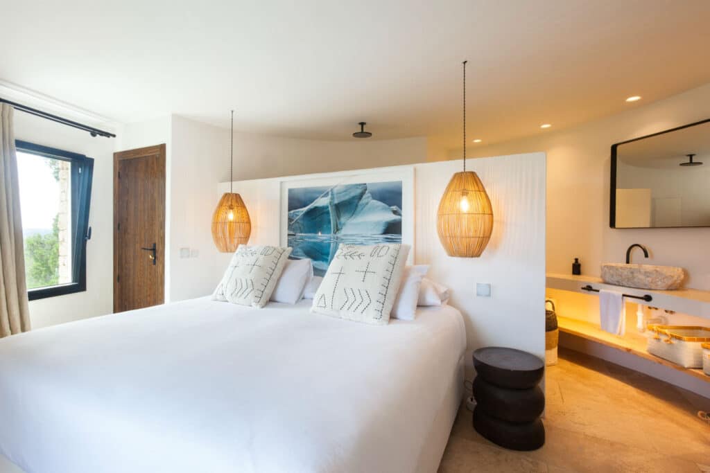 A bright, modern bedroom evokes the feel of luxury travel with a large bed adorned in white linens and decorative pillows. Two wicker pendant lights hang elegantly above. A large window invites natural light, while a photograph of a wave graces the wall above the bed. David Pearson Travel