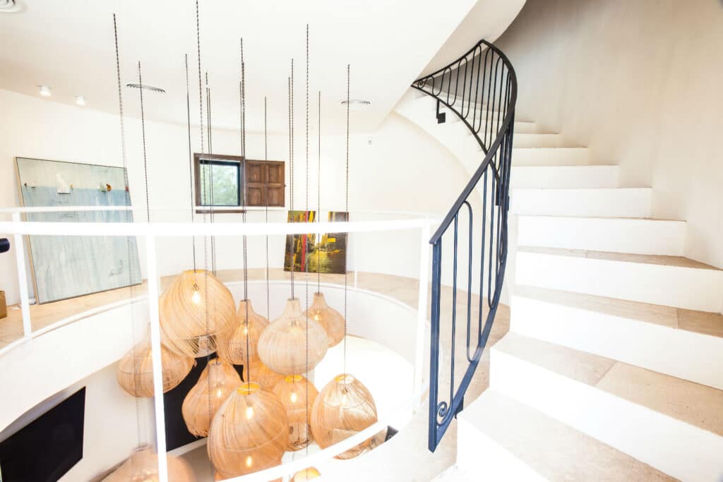 A bright, modern interior exuding luxury travel vibes features a white spiral staircase with black railing on the right. Hanging wooden pendant lights illuminate the space, while a large abstract artwork adorns the wall. Light wooden flooring enhances the airy, opulent ambiance. David Pearson Travel