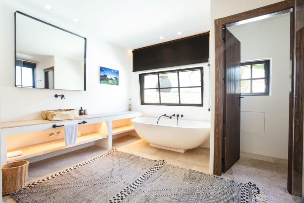 A modern bathroom evokes luxury travel with a freestanding white tub under a large window. A rectangular mirror sits above the double sink vanity with open shelves, complemented by a woven rug on the floor. The open door invites exploration, while a small painting adds a touch of elegance. David Pearson Travel