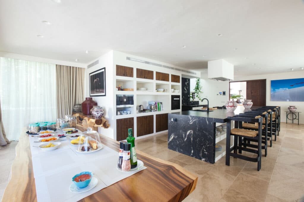 Spacious kitchen with a long, wooden breakfast bar and black stools. A black marble island features a cooktop and range hood, echoing a luxury travel experience. Shelves display ceramics, while a table holds breakfast items, drinks, and condiments. Large windows allow ample natural light. David Pearson Travel