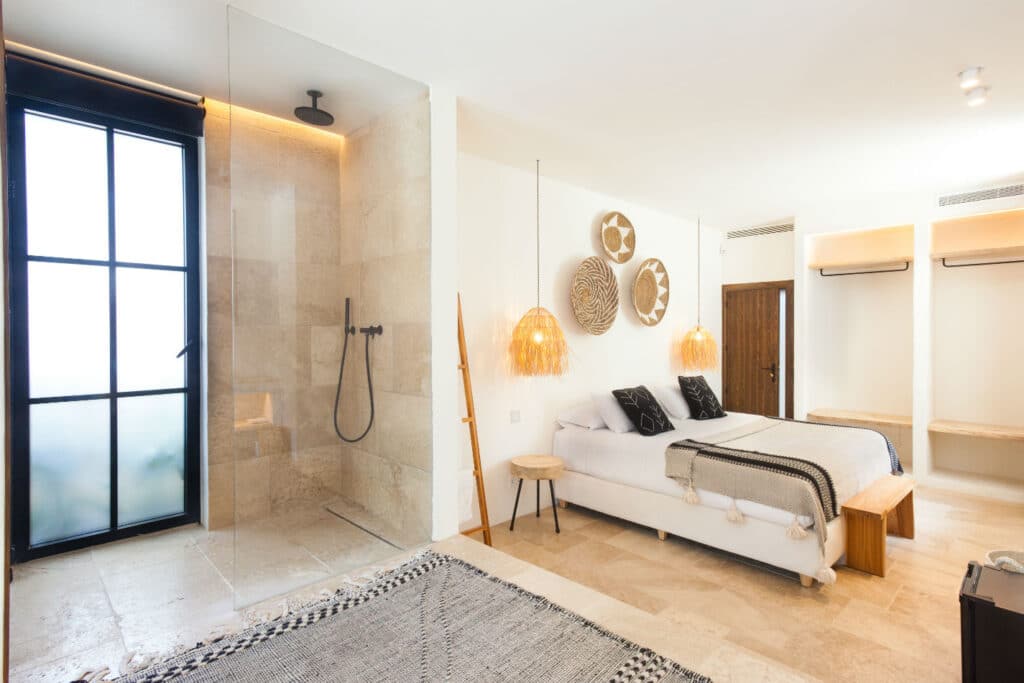 A modern bedroom exuding luxury travel vibes, featuring a large bed with pillows, decorative woven wall hangings, and hanging lamps. Adjacent is an open glass shower with beige tiles. The room boasts light-colored floors and a partially open closet with wooden shelves, perfect for the savvy traveler. David Pearson Travel
