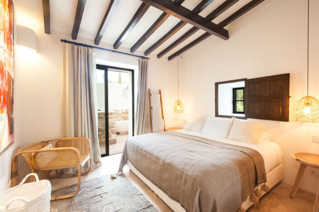 A cozy bedroom perfect for luxury travel features a large bed with beige linens, two hanging woven lamps, a small wooden chair, and a window with shutters. A door leads to an outdoor area with a stone wall, while wooden ceiling beams add warmth and sophistication to the room. David Pearson Travel