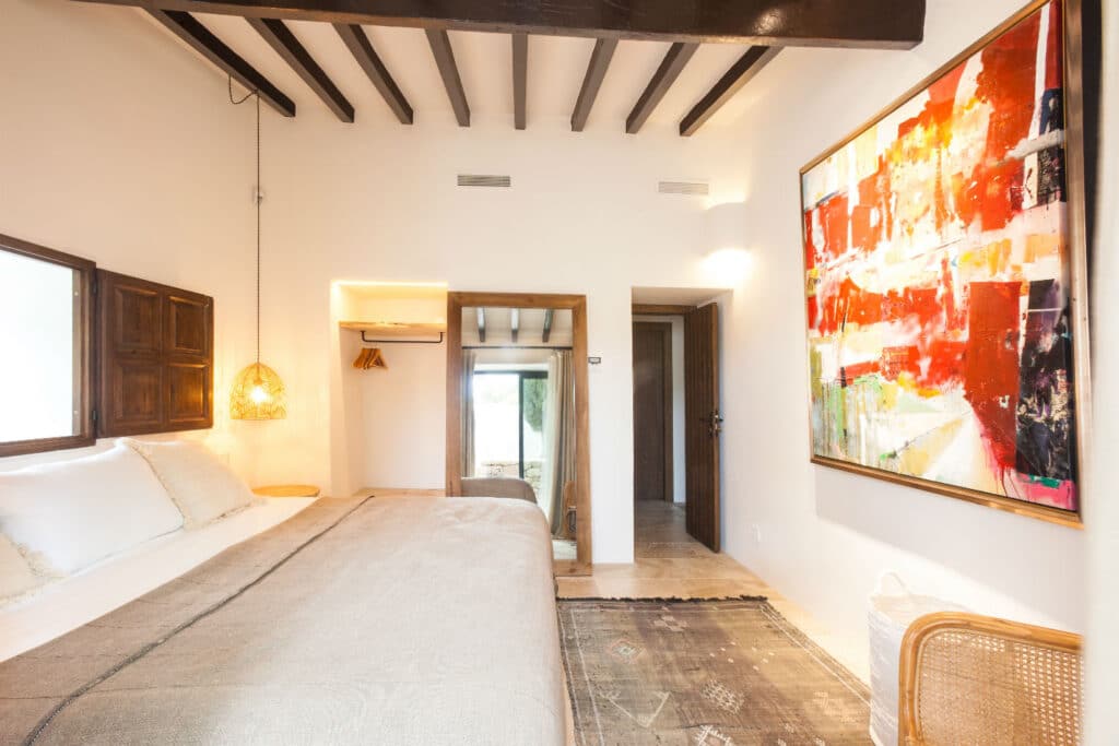 A bright bedroom with a large bed covered in neutral linens offers a touch of luxury travel appeal. Sunlight streams in from windows, illuminating the modern art piece with bold red and orange hues. Exposed wooden beams and a woven hanging lamp add warmth to this elegant getaway. David Pearson Travel