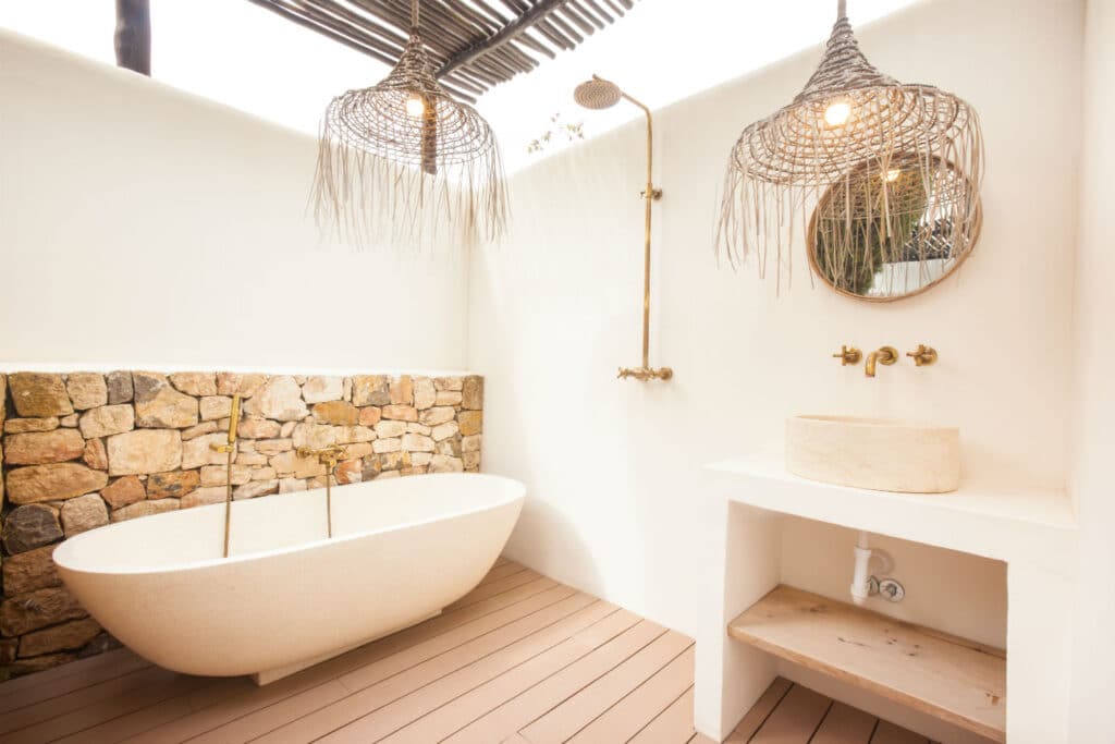 A minimalist outdoor bathroom exuding luxury travel vibes features a stone wall, freestanding oval bathtub, wooden floor, and round mirror above a stone sink. Brass fixtures and wicker light fixtures complete the rustic design for a serene escape. David Pearson Travel