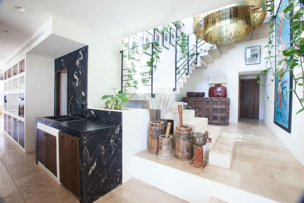 Modern interior with a marble-topped sink, wooden accents, and potted plants evokes the allure of luxury travel. A staircase adorned with hanging plants leads to a landing with framed art. A large, round, golden chandelier hangs prominently above the stone tile flooring that completes the look. David Pearson Travel