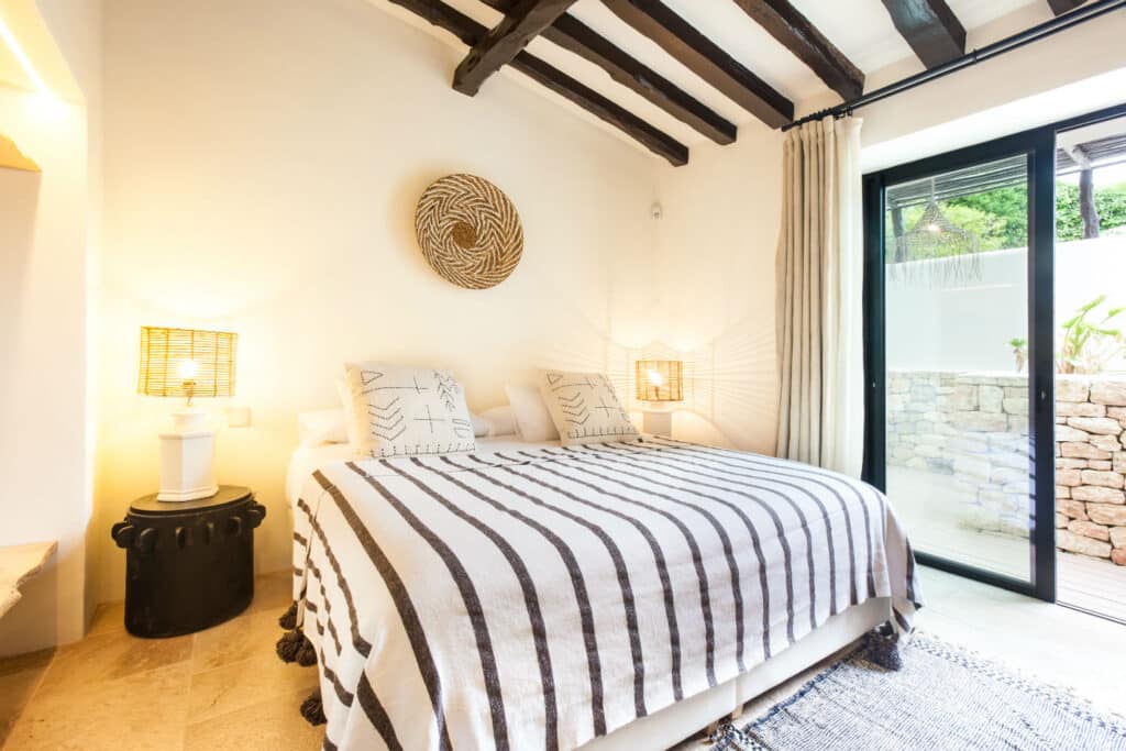 A cozy bedroom exudes a luxury travel vibe with a bed adorned by a striped blanket, two patterned pillows, and a decorative woven piece on the wall. Two lamps with textured shades rest on small bedside tables. A sliding glass door reveals a patio with an elegant stone wall. David Pearson Travel