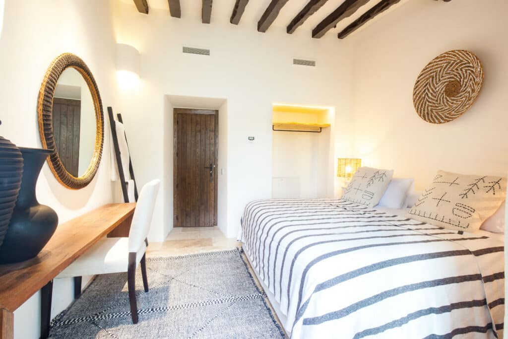 A cozy bedroom with a rustic wooden ceiling and beams exudes luxury travel vibes, featuring a large bed with striped bedding. A round mirror and decorative woven basket adorn the walls, while a wooden desk and chair sit beside the bed on a patterned rug. David Pearson Travel