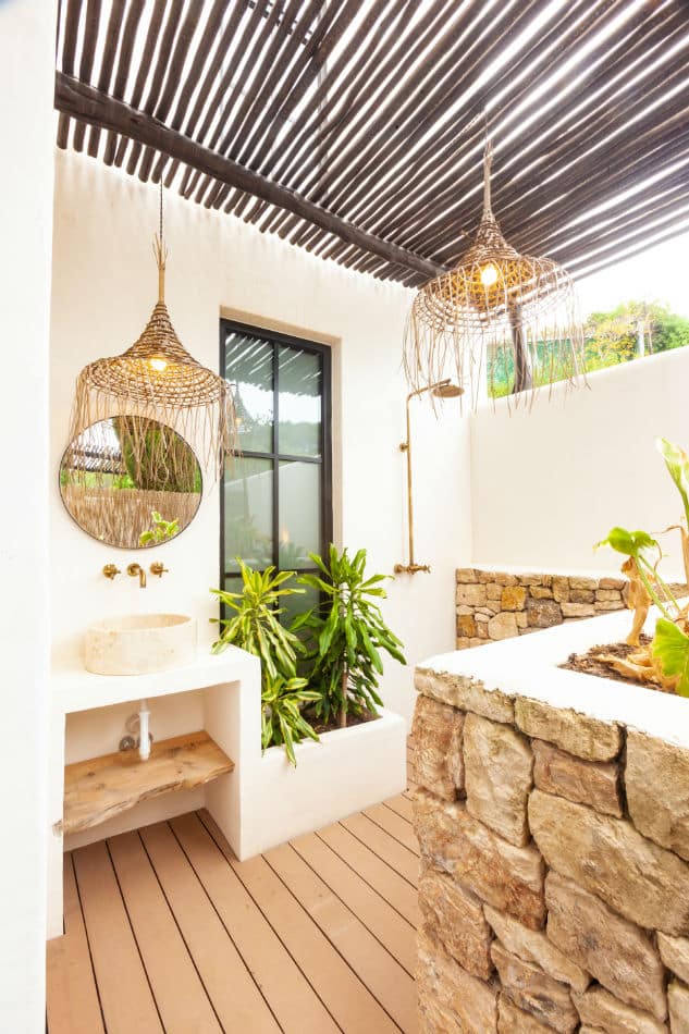 This luxurious outdoor bathroom boasts wooden flooring and a stone wall, enhanced by a round mirror and wicker lampshades. A concrete sink and rain shower offer indulgence, while potted plants add greenery. The pergola with slats allows sunlight to filter through, creating an exquisite luxury travel experience. David Pearson Travel