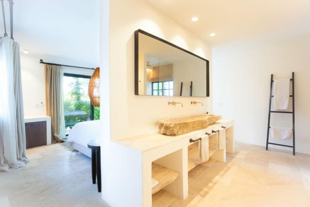 A modern bathroom exudes luxury with a large rectangular mirror above a stone sink and wooden countertop. Twin faucets enhance the elegance, while a towel ladder and beige tiles complete the bright, minimalist decor. A bed peeks from the adjacent room, promising comfort akin to luxury travel. David Pearson Travel