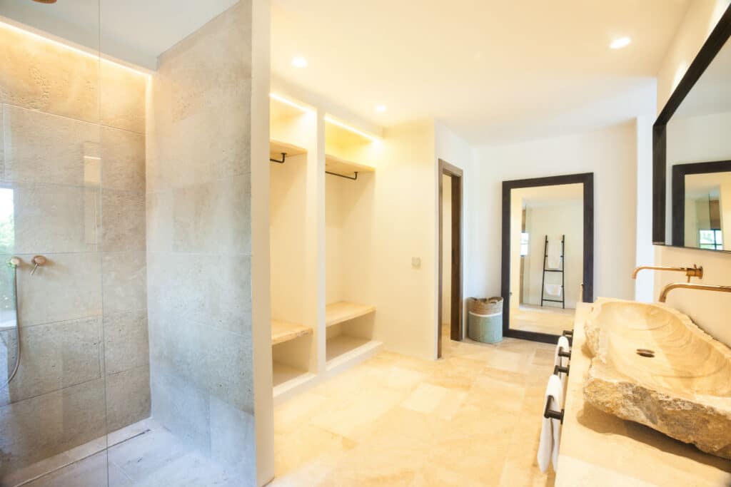 Experience luxury travel right at home in this spacious bathroom featuring a large stone sink with bronze fixtures, an open shower adorned with beige tiles, built-in wardrobes, and a large mirror. Recessed lighting and natural light from the window enhance the opulent ambiance. David Pearson Travel