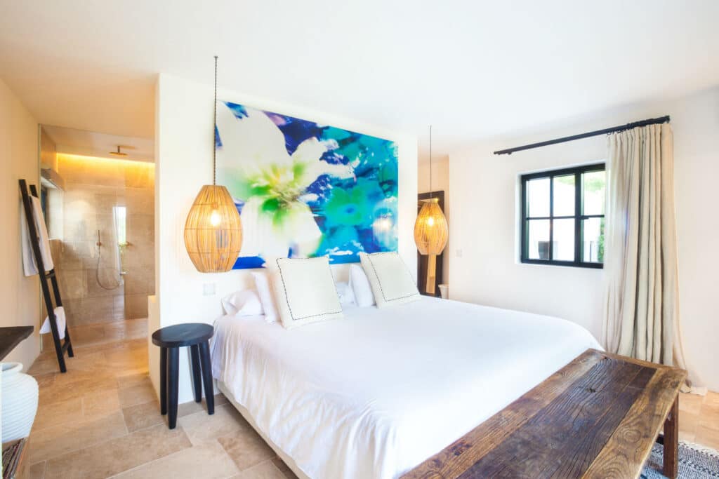 A bright bedroom epitomizing luxury travel features a large bed with white linens and three pillows. Decorative hanging lights frame a floral mural above the bed. The room flaunts light tile flooring, a wooden bench, and an open door to the bathroom. A window with curtains is on the right. David Pearson Travel