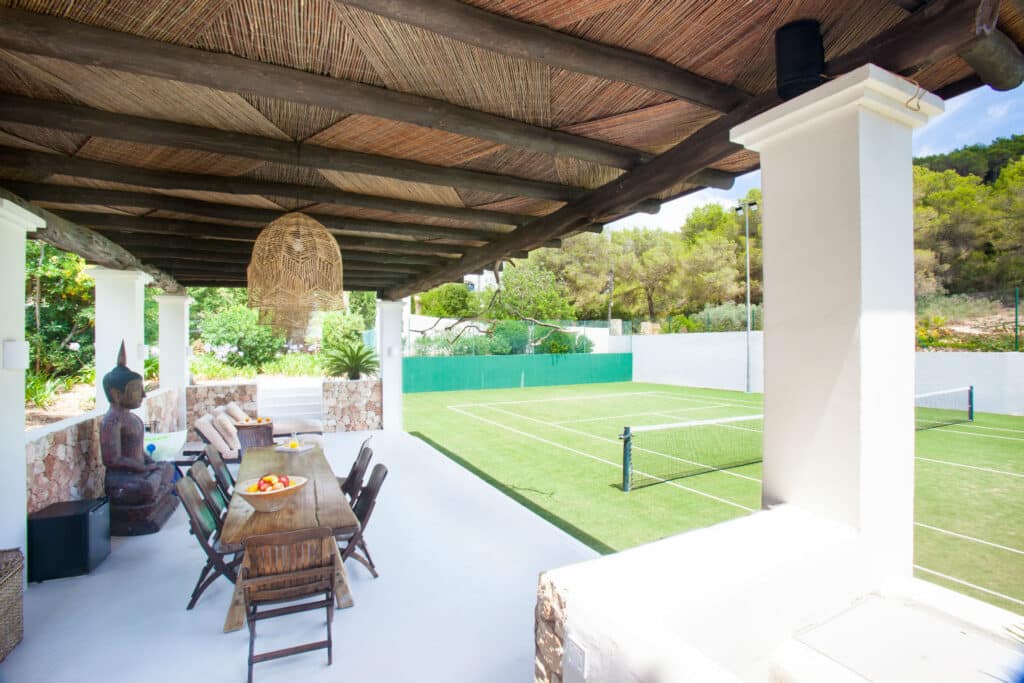 Experience luxury travel at its finest with a covered outdoor dining area featuring a wooden table and wicker chairs, right next to the tennis court. This serene space boasts a thatched roof, decorative lighting, and a large statue, all enveloped by lush greenery for an unparalleled ambiance. David Pearson Travel