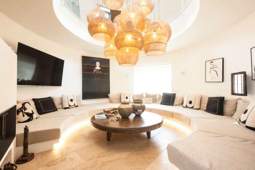 A cozy living room with a circular seating arrangement and a wooden coffee table at the center exudes luxury. Above, wicker pendant lights hang from the ceiling. The walls are adorned with minimalist art, evoking the elegance of luxury travel, and a large TV is mounted on the wall. David Pearson Travel