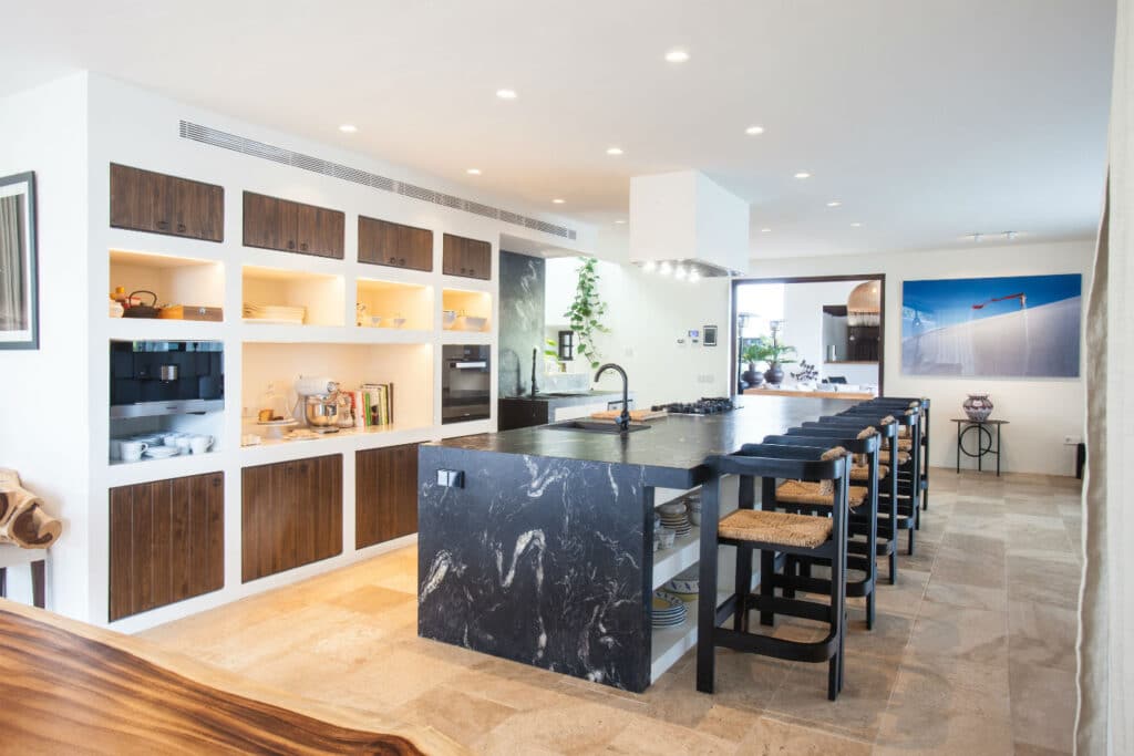 A modern kitchen with an island featuring a black marble countertop and bar stools offers a luxury travel vibe. Open shelving with dishes, built-in ovens, and wooden cabinets are on the left. The well-lit space features recessed lighting and tiled flooring, perfect for culinary adventures. David Pearson Travel