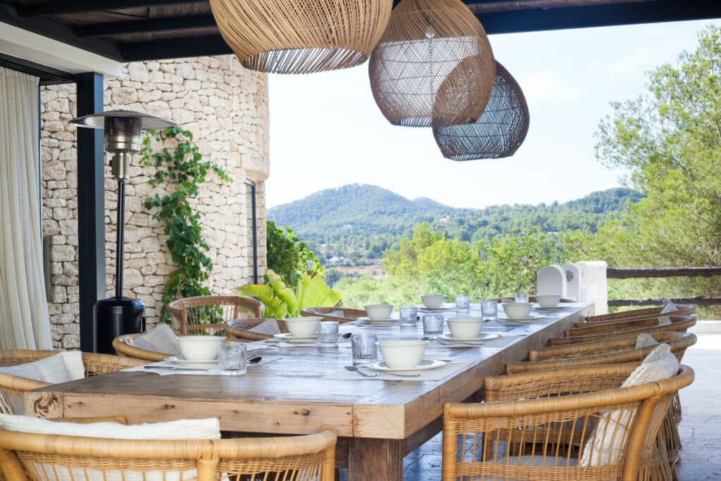 A rustic outdoor dining area with a long wooden table set for a meal exudes the charm of luxury travel. Wicker chairs surround the table, and large wicker pendant lights hang above, all framed by a view of greenery and hills in the background. David Pearson Travel