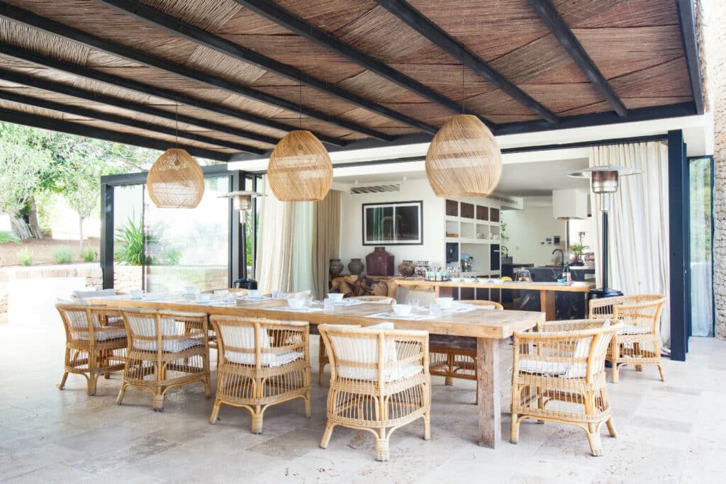 Experience luxury travel with a spacious outdoor dining area featuring a long wooden table and wicker chairs under a thatched roof. Three large wicker pendant lights hang overhead, complementing the open kitchen with modern appliances and expansive sliding glass doors. David Pearson Travel