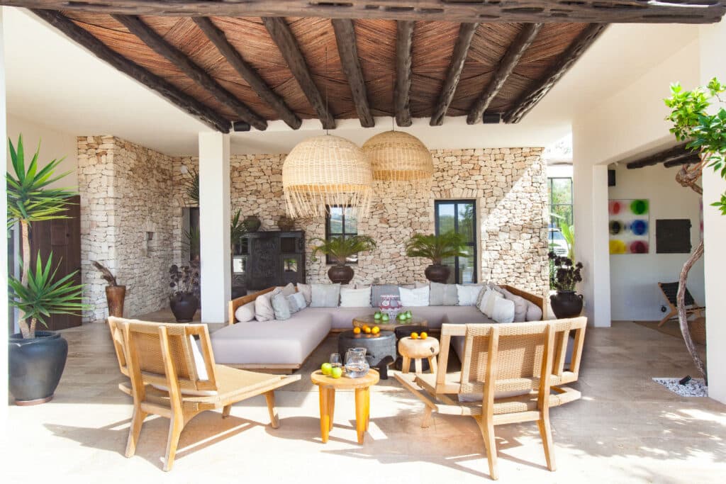 A cozy patio designed for luxury travel enthusiasts, featuring a large L-shaped sofa and two wooden chairs centered around a low table adorned with fruit. Stone walls, woven ceiling details, large pendant lights, and vibrant potted plants create a serene atmosphere. David Pearson Travel
