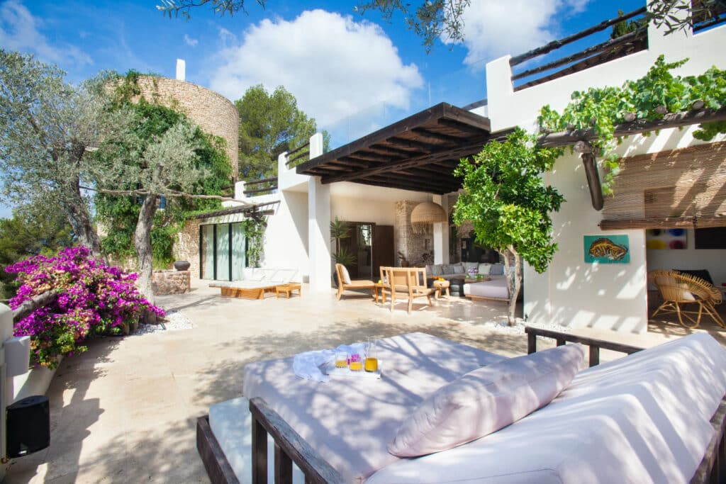 Experience luxury travel on the sunny outdoor patio of a Mediterranean villa, featuring white walls and wooden accents. Relax on a cushioned lounge bed surrounded by vibrant pink bougainvillea and potted plants, with shaded seating under a pergola against a backdrop of trees and clear blue sky. David Pearson Travel