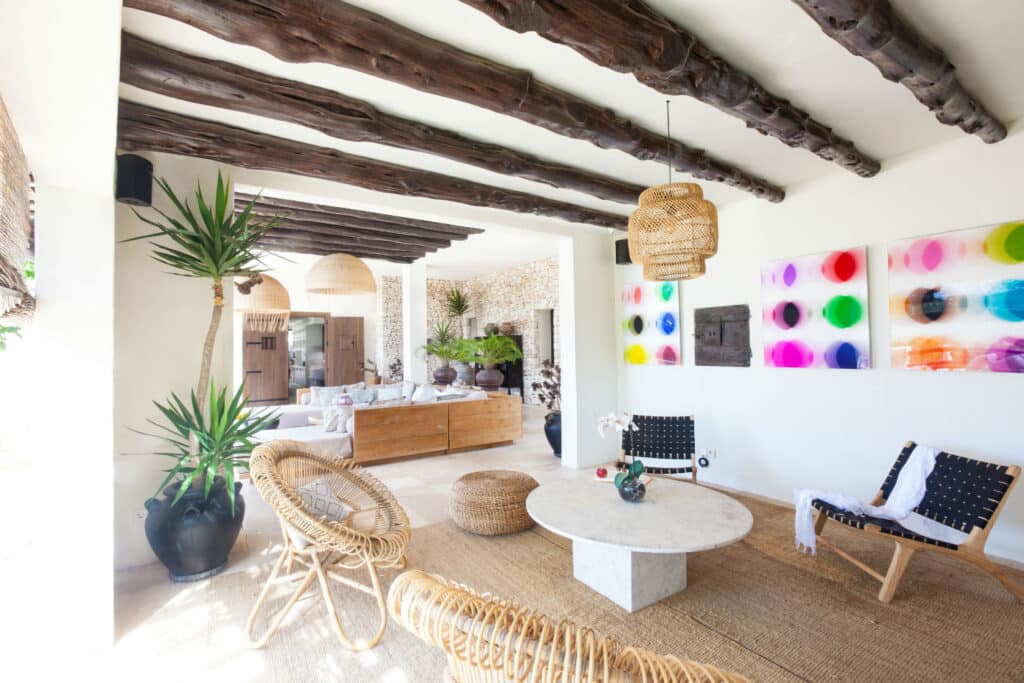 Discover a spacious living room that evokes the essence of luxury travel, featuring exposed wooden beams and natural decor. Enjoy wicker chairs, a marble coffee table, vibrant abstract artwork, and large potted plants that together create an effortlessly bright, airy atmosphere. David Pearson Travel