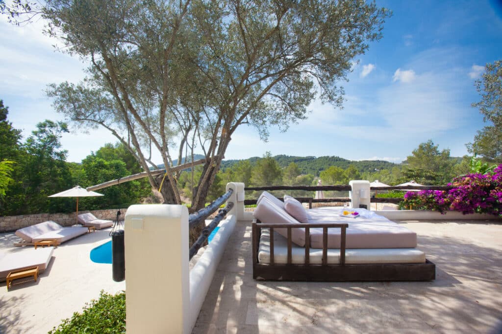 A spacious outdoor terrace epitomizes luxury travel with a large daybed overlooking lush greenery and a small swimming pool below. Sun loungers and a parasol sit by the pool, while vibrant purple flowers adorn the edge of the terrace under a clear blue sky. David Pearson Travel