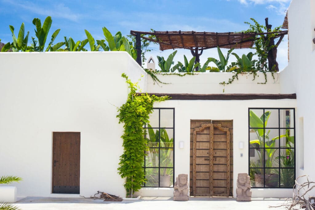 A white building with a wooden door and two statues on either side reflects luxury travel charm. Large windows flank the door, green vines climb the walls, and a terrace with a rustic pergola and lush greenery decorates the rooftop beneath the clear blue sky. David Pearson Travel