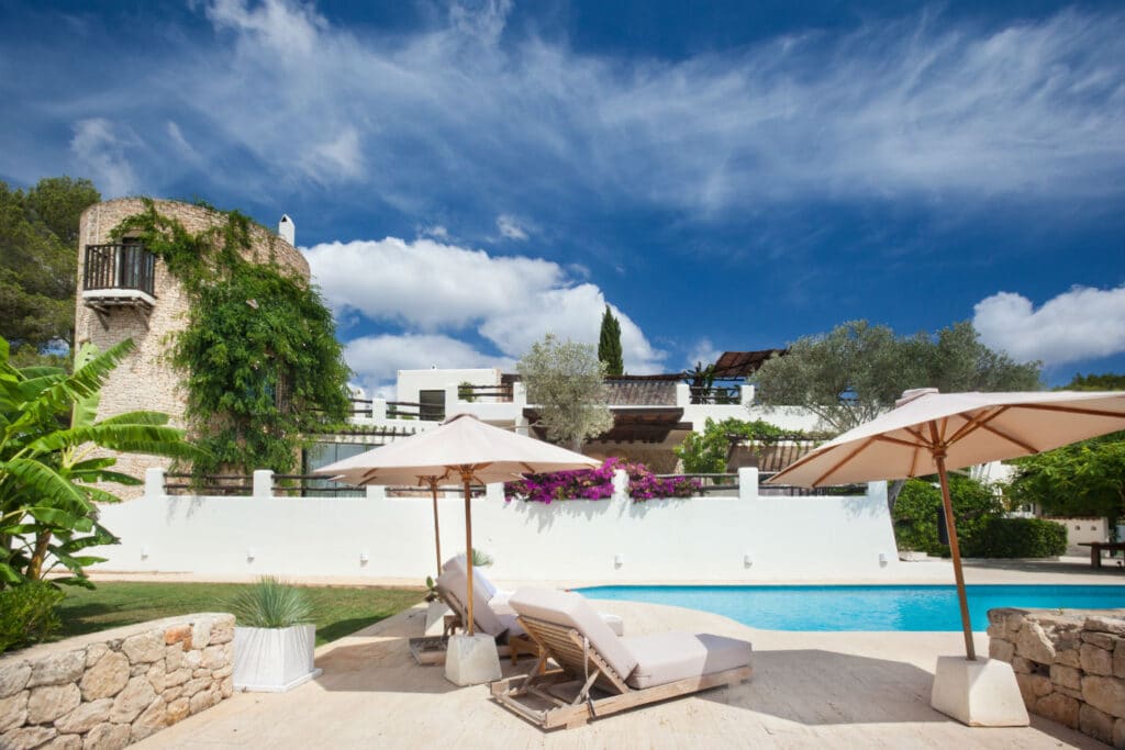 A luxurious villa with white walls and a stone tower overlooks a clear blue swimming pool, embodying the essence of luxury travel. Two lounge chairs with umbrellas are positioned by the pool, while lush greenery and vibrant purple flowers enhance the serene atmosphere. David Pearson Travel