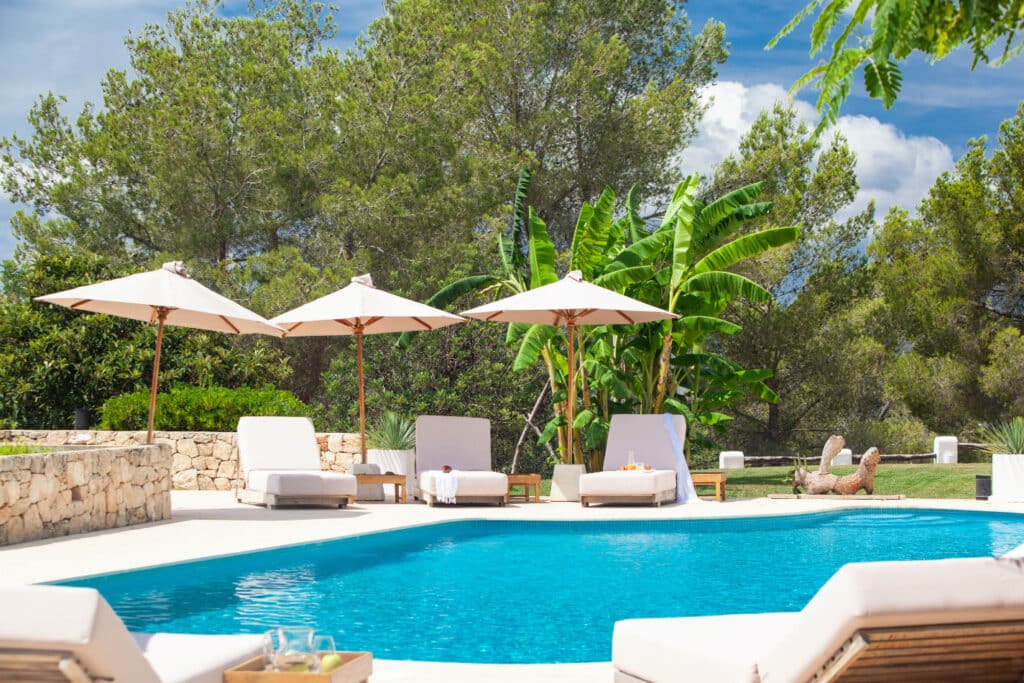 A serene outdoor pool area epitomizing luxury travel, with lounge chairs and umbrellas set around a clear blue pool. Lush greenery, including tall trees and banana plants, surrounds the space under a bright blue sky. A touched wooden tray with drinks rests at the poolside edge. David Pearson Travel