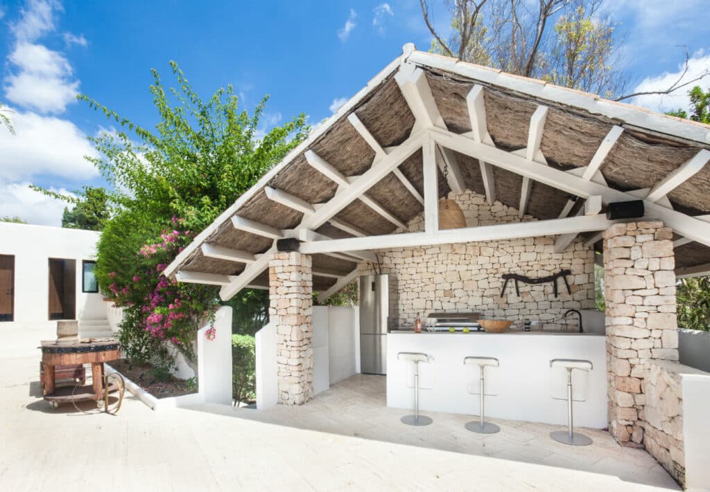 An outdoor kitchen with a thatched roof and stone walls exudes the charm of luxury travel, featuring a bar counter with three modern stools. Equipped with a mini fridge, grill, and mounted kitchen tools, its surrounded by lush greenery and set against a clear blue sky. David Pearson Travel