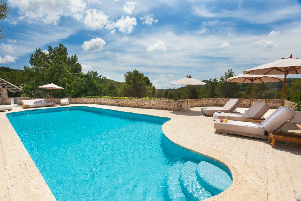 A serene outdoor pool area with turquoise water, surrounded by beige stone tiles, offers a taste of luxury travel. Lounge chairs with white cushions and umbrellas are on the right. Green trees and a partly cloudy sky create a peaceful backdrop, perfect for relaxation. David Pearson Travel