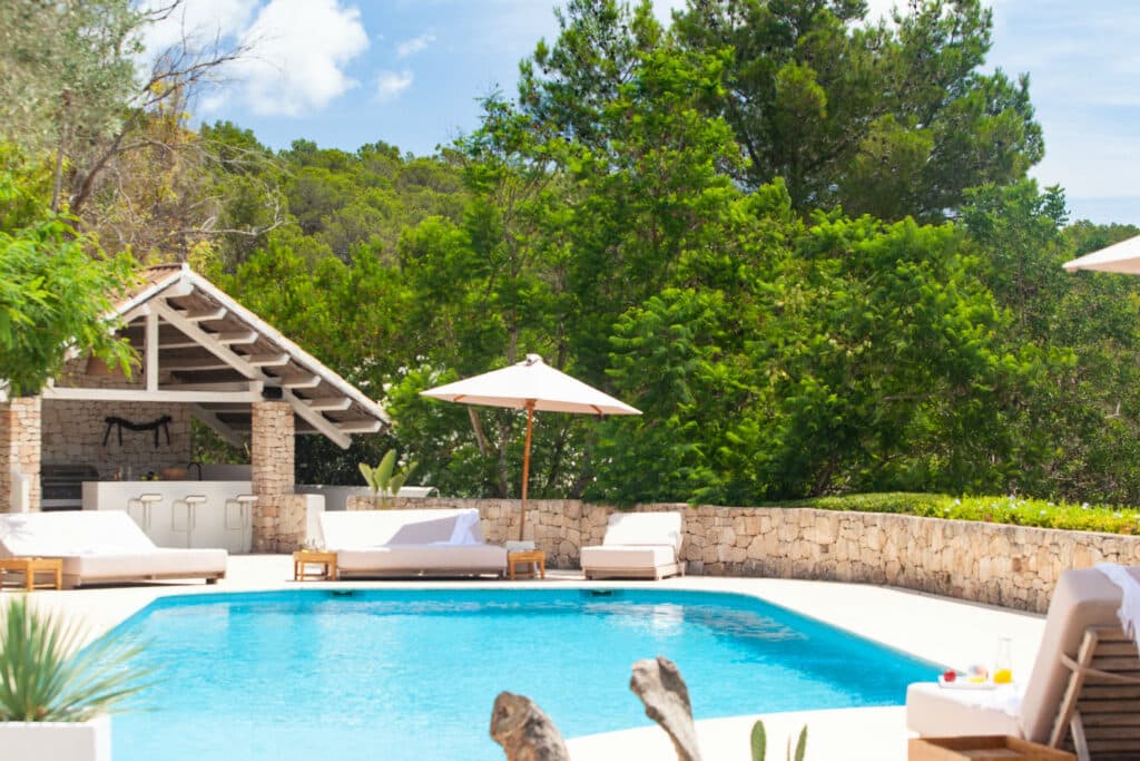 Experience the essence of luxury travel with a sumptuous pool embraced by stone walls and lush greenery. Lounge chairs with white cushions and umbrellas encircle the water, while a rustic, open-air cabana features a bar area in the background beneath a partly cloudy sky. David Pearson Travel