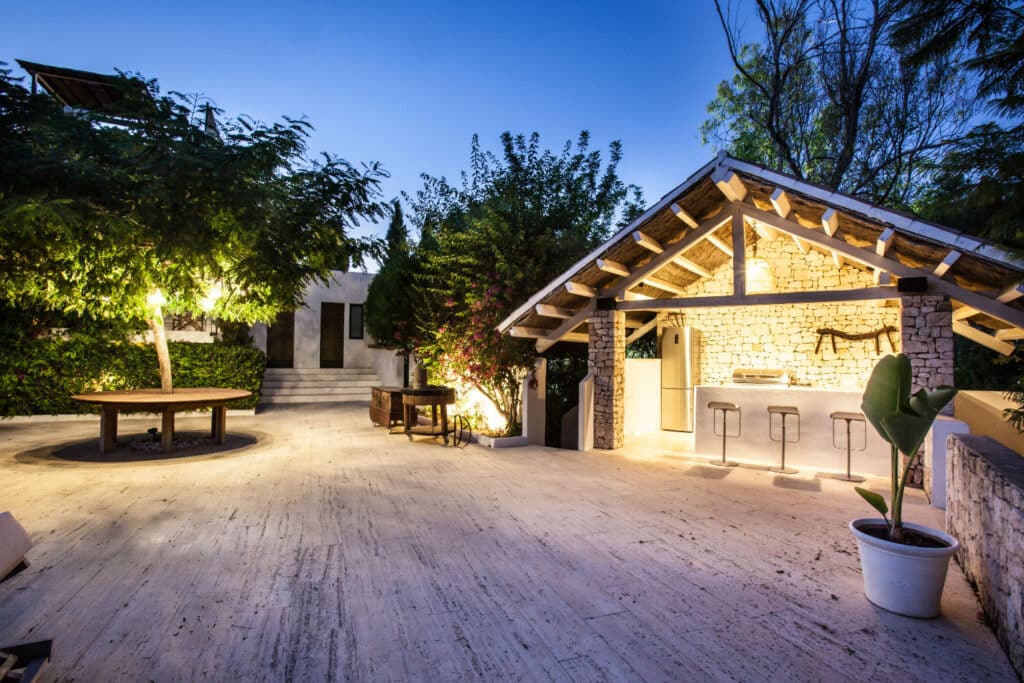 The outdoor patio area at dusk exudes luxury travel, featuring a stone and wood pavilion with a bar and three stools. Lush trees and plants envelop the space, while a round table sits to the left. Steps lead invitingly to a pristine white building in the background. David Pearson Travel