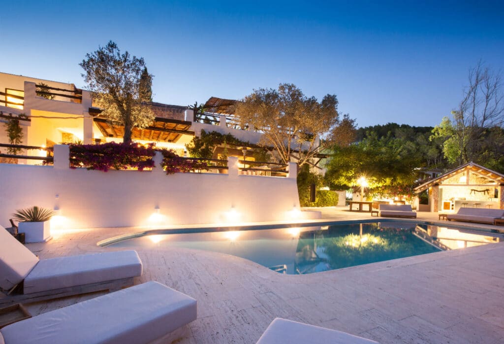 A luxurious villa perfect for luxury travel, with a lit swimming pool in the foreground, surrounded by white walls and lush greenery. Loungers encircle the pool, and a pergola draped in climbing plants stands silhouetted against the twilight sky. David Pearson Travel
