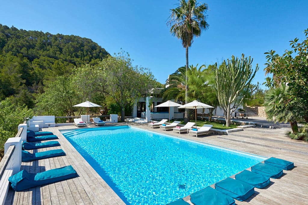 A bright outdoor pool with crystal-clear water is surrounded by wooden decking and lined with blue sun loungers, offering a luxury travel experience. White umbrellas and lounge chairs dot the area, while lush greenery and palm trees create a serene backdrop under the clear blue sky. David Pearson Travel