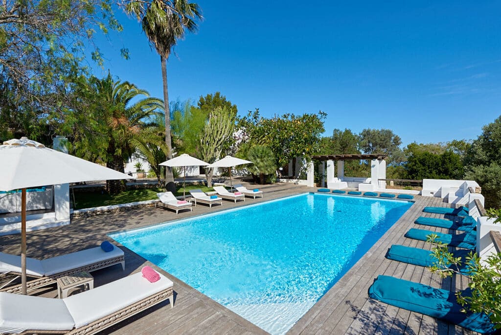 Experience the epitome of luxury travel at a serene outdoor pool area surrounded by lush greenery and palm trees. Lounge chairs with white and blue cushions line the wooden deck, while parasol umbrellas provide shade, crafting a relaxing atmosphere under a clear blue sky. David Pearson Travel