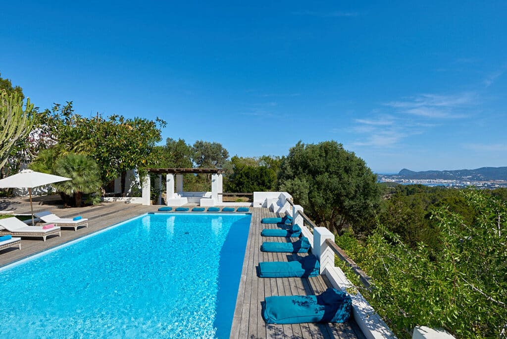 A scenic outdoor pool with turquoise water is lined with blue sun loungers, offering the perfect luxury travel experience. Surrounded by lush greenery, a white pergola draped in leafy vines provides shade, while distant hills and a clear blue sky complete this idyllic setting. David Pearson Travel