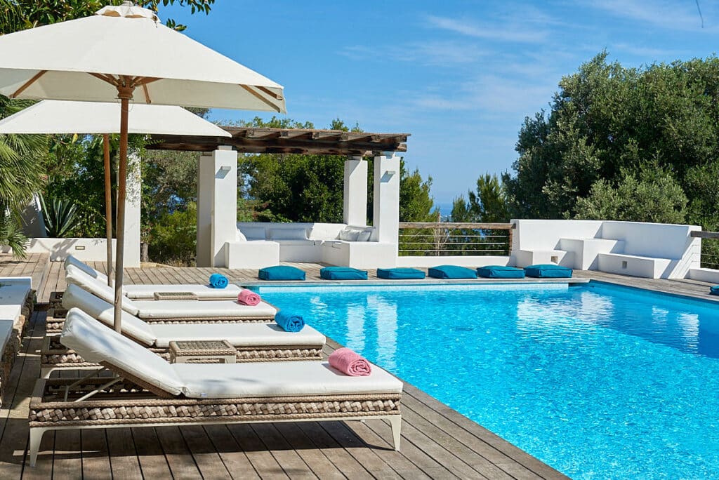 Luxury travel awaits with an outdoor pool area featuring several white lounge chairs, each topped with a rolled towel. A large umbrella provides shade, adding to the opulent experience. In the background, a wooden pergola shelters a seating area as clear water reflects the sunny sky. David Pearson Travel