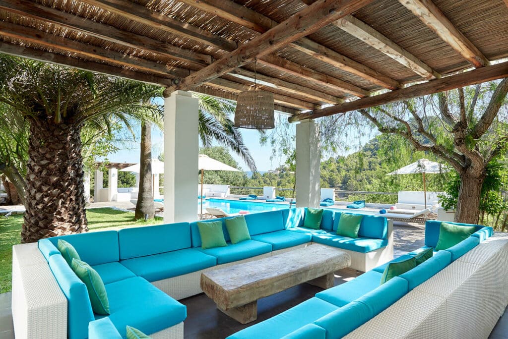This luxury travel oasis features an outdoor lounge with white wicker sofas adorned with bright turquoise cushions, arranged in a large U-shape around a rustic wooden table. A wooden pergola provides shade, while the backdrop of a pool, palm trees, and distant greenery completes the idyllic scene. David Pearson Travel